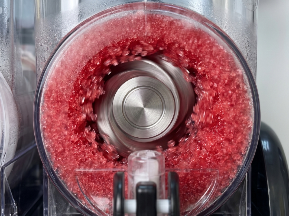 A close-up view of a metal auger spinning inside a clear slushy machine bowl, actively churning bright red icy mixture to maintain a consistent frozen texture.