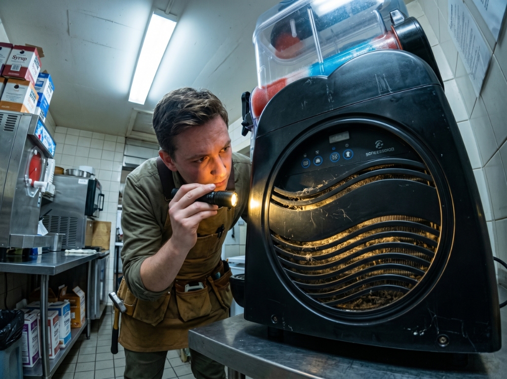 A male maintenance technician in a brown apron uses a bright flashlight to inspect a heavily clogged and dusty condenser grill on the side of a commercial slush machine.