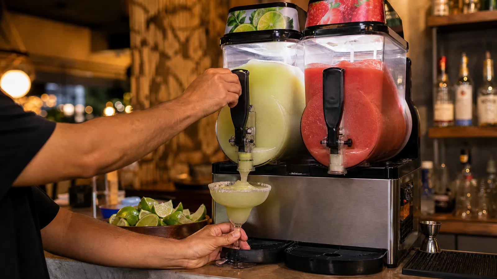 Bartender pulling the tap of a two-bowl frozen drink machine to fill a salt-rimmed glass with frozen margarita, with a red strawberry slush in the second bowl and lime wedges on the bar